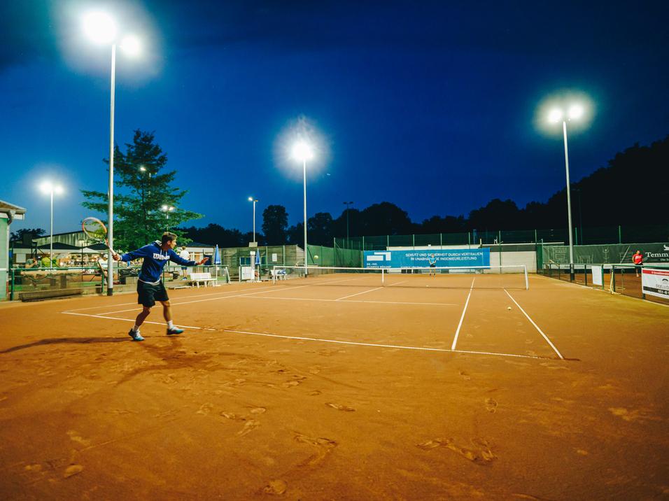 Tennisspieler schlägt in der Dämmerung auf einem beleuchteten Sandplatz.