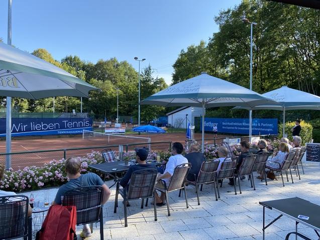 Blick auf einen Tennisplatz mit Zuschauern in Stühlen unter Schirmen, umgeben von Blumenbeeten und Bäumen.