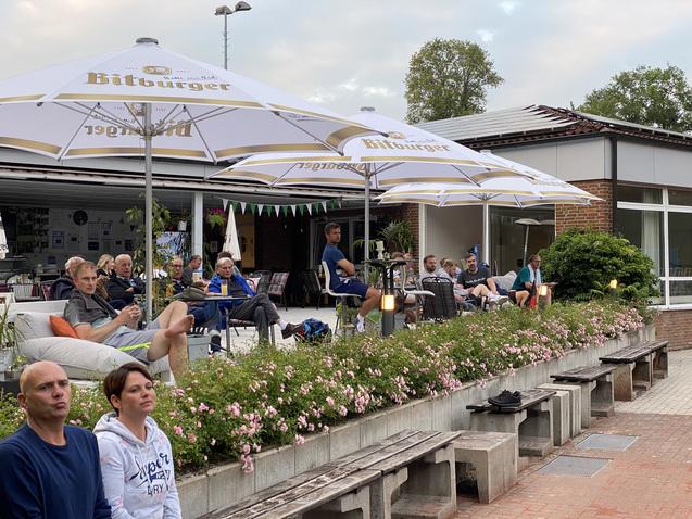 Gruppe von Menschen sitzt unter Schirmen an einem Café im Freien, umgeben von Blumen und einer entspannenden Atmosphäre.
