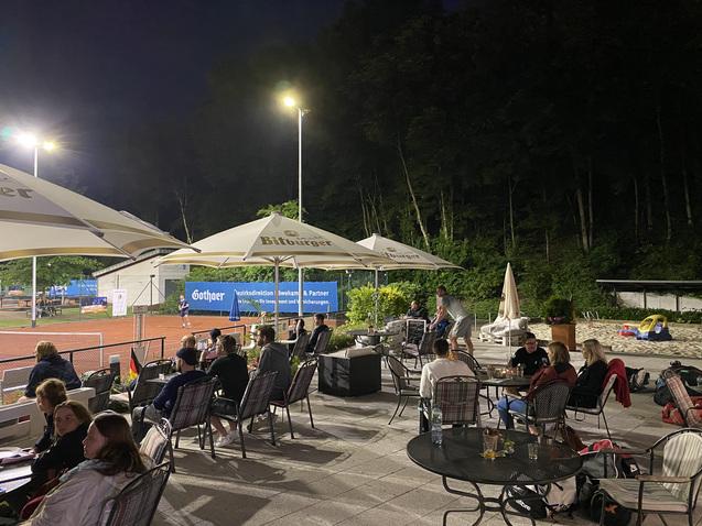 Personen sitzen abends an Tischen unter Sonnenschirmen neben einem Tennisplatz, umgeben von Bäumen und Beleuchtung.