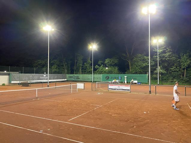 Tennisplatz bei Nacht mit Beleuchtung, zwei Spielern und Werbebannern im Hintergrund. Rote Sandoberfläche und grüne Umgebung.