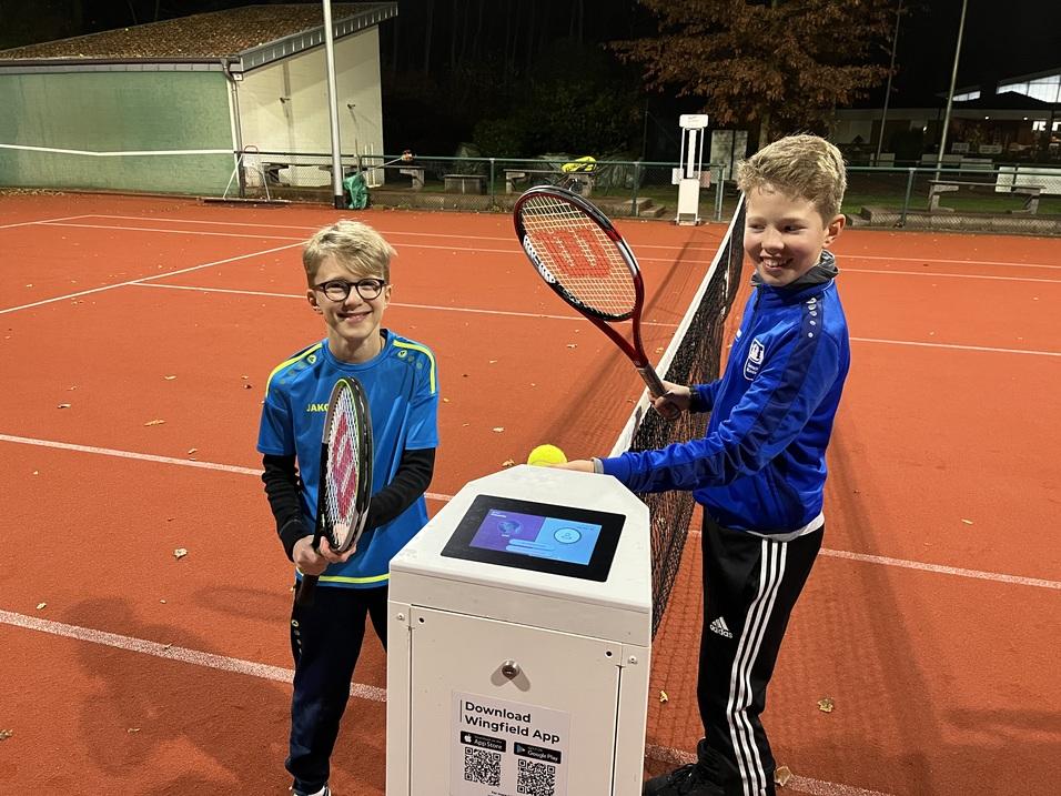 Zwei Kinder halten Tennisschläger und stehen an einem Infostand auf einem beleuchteten Tennisplatz.