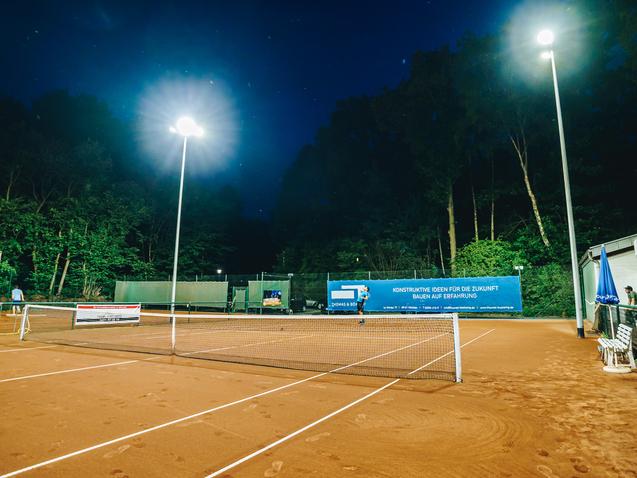 Beleuchteter Tennisplatz mit roten Sand, Bänken und Werbebanner unter einem nächtlichen Himmel.