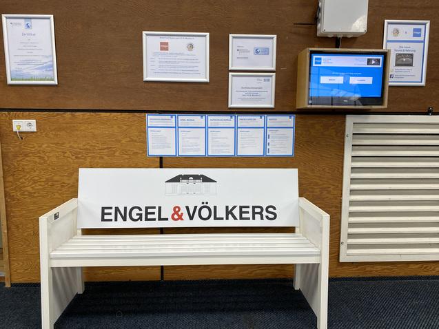 Weißer Informationsstand mit der Aufschrift „ENGEL & VÖLKERS“ und mehreren Zertifikaten an der Wand.