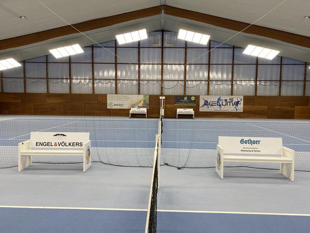 Blick in eine Tennishalle mit zwei Bänken und Netzen auf einem blauen Platz, umgeben von Werbebannern.