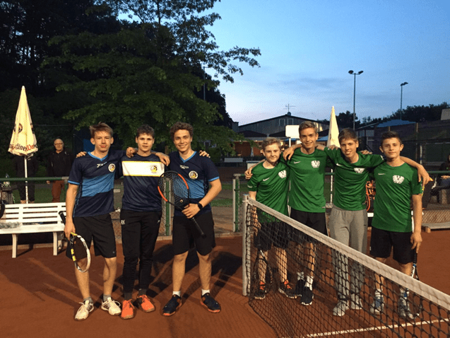 Gruppe von sieben Jugendlichen in Sportkleidung, lächelnd auf einem Tennisplatz bei Abenddämmerung, einige mit Schlägern.