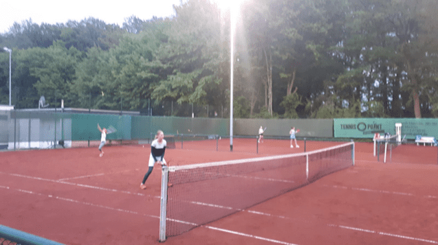 Zwei Tennisspielerinnen in Aktion auf einem Tennisplatz mit roten Sandboden, umgeben von Bäumen bei Abendlicht.