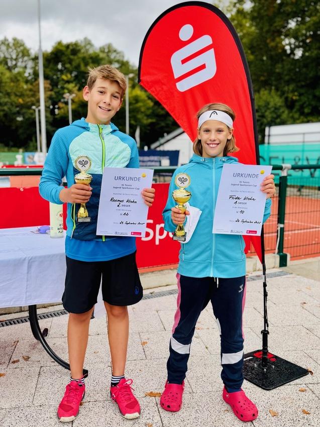 Zwei Kinder stehen mit Pokalen und Urkunden auf einem Sportplatz, im Hintergrund ein Banner und Tennisplätze.