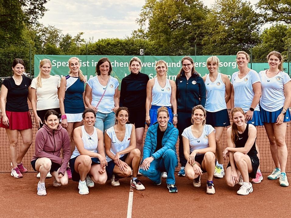 Gruppe von 16 Frauen in Sportkleidung posiert auf einem Tennisplatz mit Bäumen im Hintergrund.