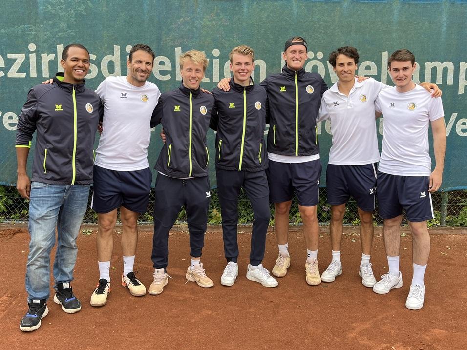 Gruppenfoto von acht Männern in Sportkleidung auf einem Tennisplatz, im Hintergrund eine Werbetafel.