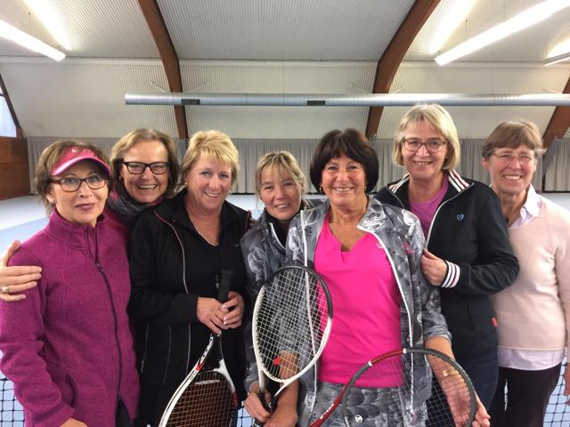 Gruppe von sieben Frauen in Sportkleidung mit Tennisschlägern, lächelnd in einer Tennishalle posierend.