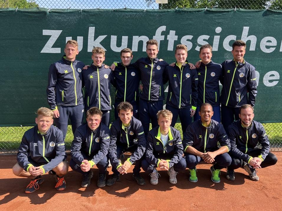 Gruppe von 14 jungen Tennis-Spielern in Sportjacken, lächelnd vor einem grünen Netz auf einem Tennisplatz.