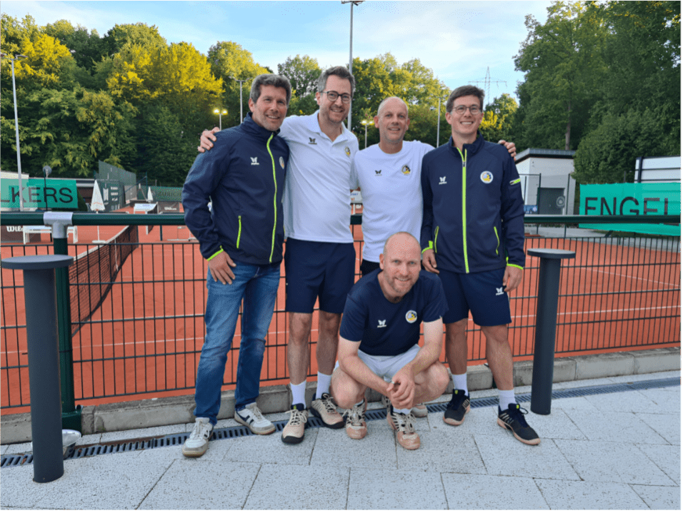 Fünf Männer posieren lächelnd vor einer Tennisanlage mit orangefarbenem Belag und grüner Umzäunung im Hintergrund.