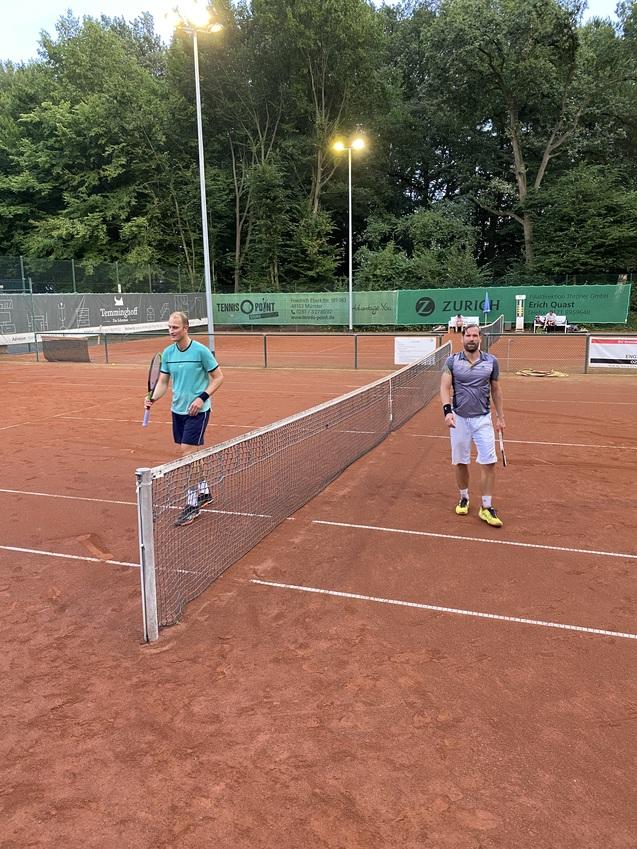 Zwei Tennisspieler stehen an einem Netz auf einem roten Tennisplatz, umgeben von Bäumen und Lichtern.