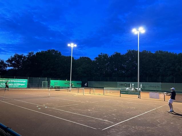 Tennisplatz bei Dämmerung mit Flutlicht und einem Spieler, der sich zur Schlagposition vorbereitet.