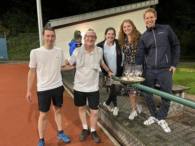 Fünf Personen stehen auf einem Tennisplatz und halten Gläser mit Getränken neben einem Tisch mit Snacks.