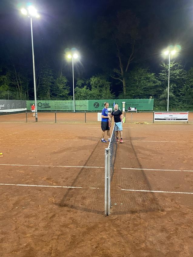 Zwei Personen stehen sich an einem Tennisnetz auf einem beleuchteten Platz gegenüber und unterhalten sich.
