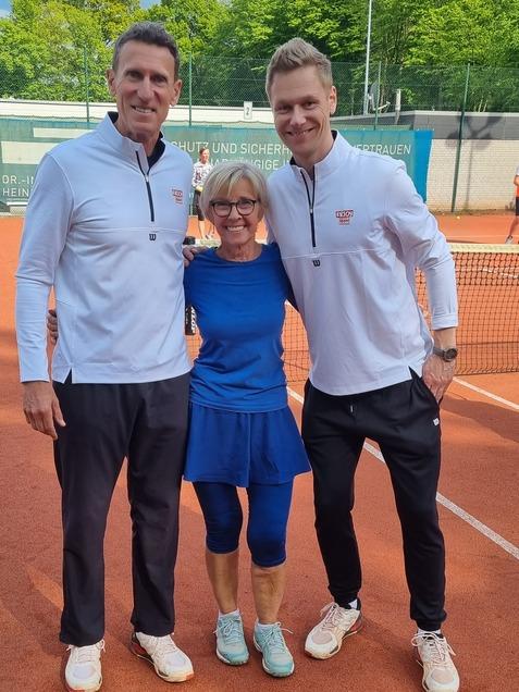 Drei Personen stehen lächelnd auf einem Tennisplatz, umgeben von grünen Bäumen und einem Tennisnetz.