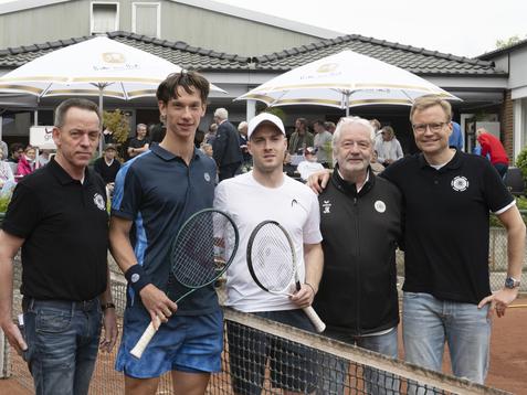 Fünf Männer stehen gemeinsam am Tennisplatz, zwei Spieler mit Schlägern, andere in sportlicher Kleidung.