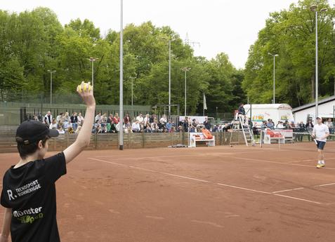 Junge wirft einen Tennisball auf einem Trainingsplatz, im Hintergrund Zuschauer auf Bänken zwischen Bäumen.