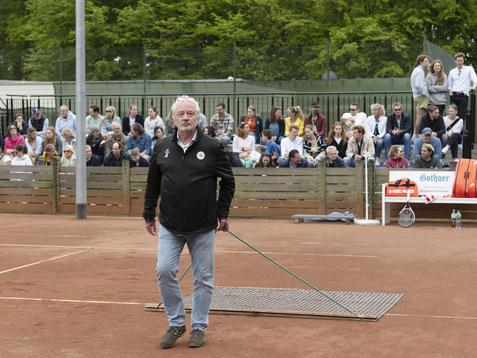 Mann mit grauem Bart zieht eine Besenplatte über einen Tennisplatz, während Zuschauer im Hintergrund beobachten.
