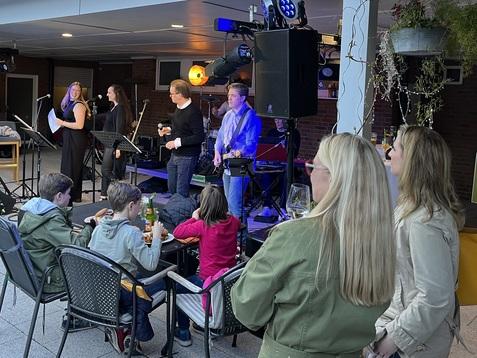 Liveband spielt im Freien, während Zuschauer in Stühlen sitzen und Getränke genießen. Beleuchtung und Pflanzen im Hintergrund.