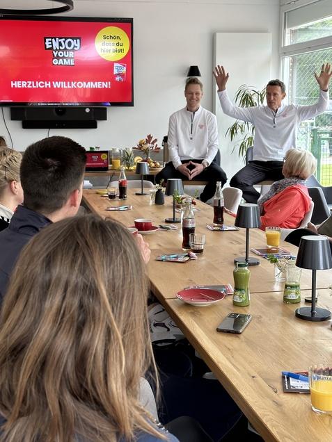Zwei Personen am Tisch halten eine Präsentation vor einer Gruppe, mit einem Bildschirm im Hintergrund, auf dem „HERZLICH WILLKOMMEN!“ steht.