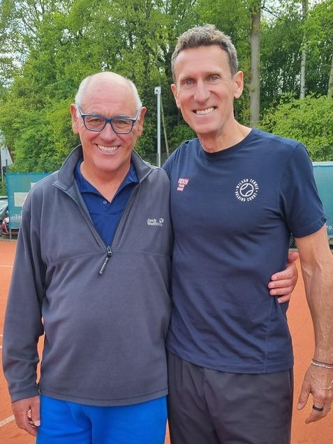Zwei Männer stehen auf einem Tennisplatz, einer trägt einen grauen Pullover und blauen Shorts, der andere ein schwarzes T-Shirt und schwarze Hose.