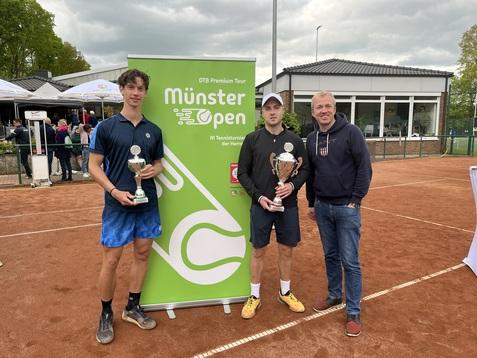 Zwei Tennis-Spieler mit Pokalen stehen neben einem Mann vor einem grünen Banner der Münster Open auf einem Sandplatz.