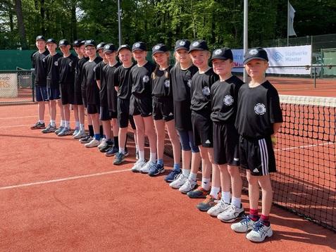 Gruppe von 15 Kindern in Sportkleidung steht auf einem Tennisplatz, umgeben von Bäumen und Sonnenschein.