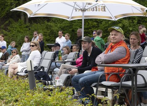 Gruppe von Zuschauern auf Stühlen unter Sonnenschirmen, während sie ein Event im Freien beobachten.