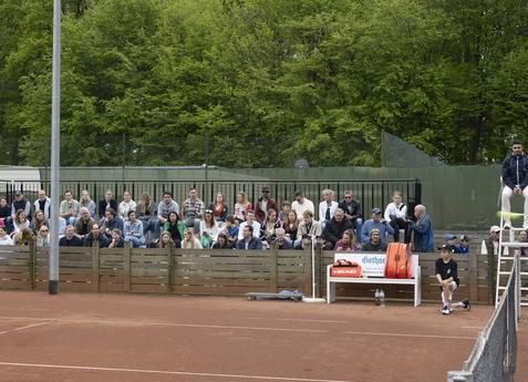Zuschauer sitzen an einem Tennisplatz, umgeben von Bäumen, während ein Spieler seinen Aufschlag vorbereitet.