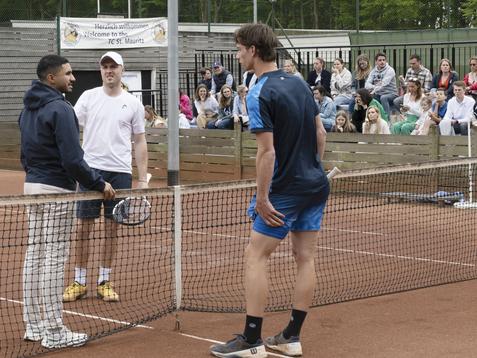 Drei Männer unterhalten sich am Tennisnetz auf einem Platz, während Zuschauer im Hintergrund zuschauen.