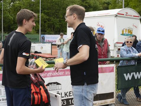Preisverleihung: Ein Jugendlicher erhält eine Auszeichnung von einem Mann auf einem Tennisplatz, Zuschauer im Hintergrund.