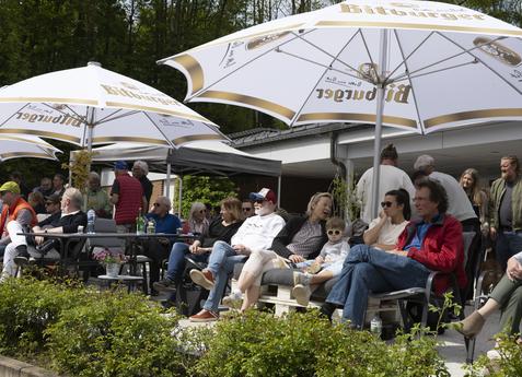 Gruppe von Menschen sitzt unter großen SonnenSchirmen in einem Garten, umgeben von Büschen und einem Gebäude im Hintergrund.