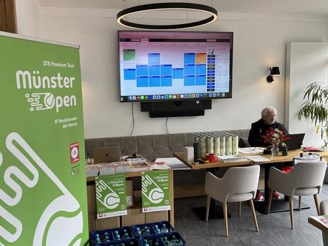 Raum mit Tisch, Monitor und Banner für das Münster Open, Person sitzt am Tisch. Getränkekisten stehen bereit.
