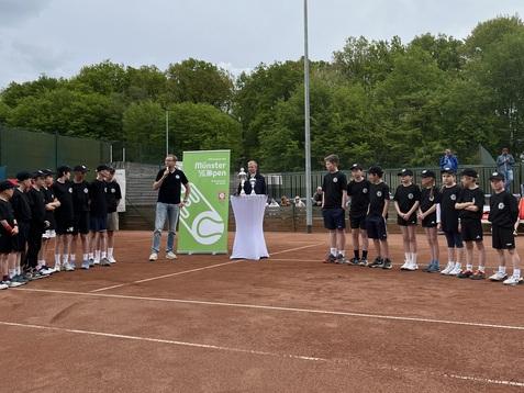 Gruppierung von Jugendlichen in Sportkleidung steht um einen Redner vor einem Tisch mit weißen Tischdecken auf einem Tennisplatz.