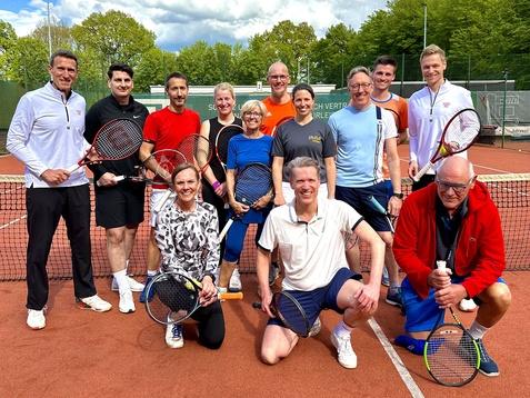 Gruppe von zwölf Tennis-Spielern posiert lächelnd auf einem Platz mit Tennisschlägern und Bällen, umgeben von Bäumen.