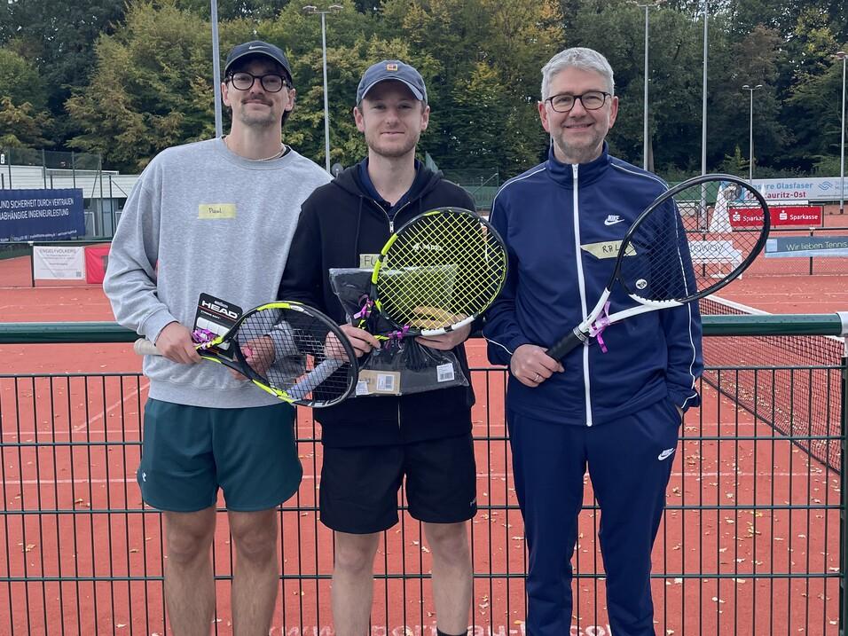 Drei Männer posieren auf einem Tennisplatz, halten Tennisschläger und Medaillen, umgeben von grünen Bäumen und Platzlichtern.