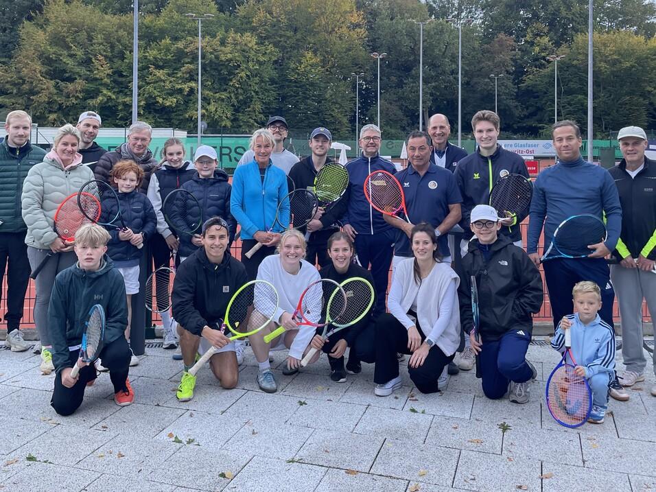 Gruppenshot von 24 Personen auf einem Tennisplatz, alle halten Tennisschläger, mit Bäumen im Hintergrund.