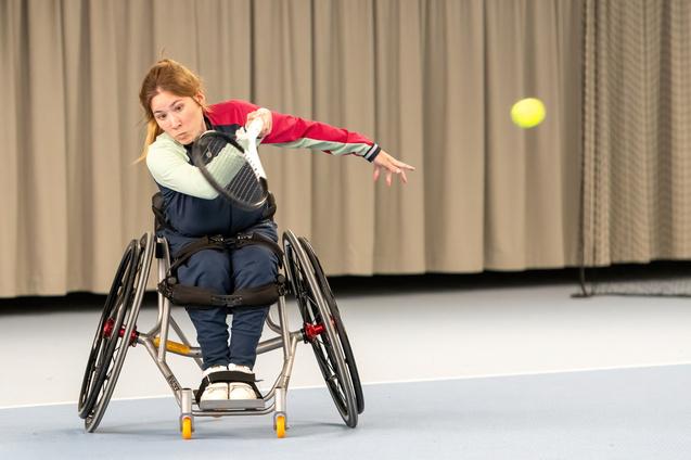Sportlerin im Rollstuhl schlägt einen Tennisschlag mit einem Schläger auf einem Indoor-Tennisplatz.