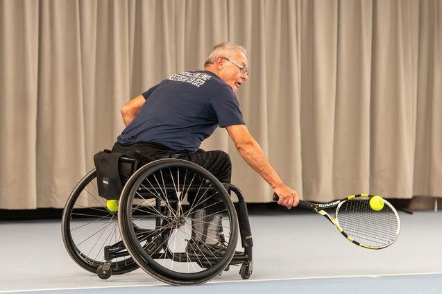 Mann im Rollstuhl schlägt mit einem Tennisracket einen Ball auf einem Tennisplatz mit grauer Rückwand.