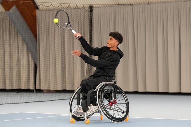 Junger Mann im Rollstuhl schlägt mit einem Tennisschläger einen Ball auf einem Indoor-Tennisplatz.