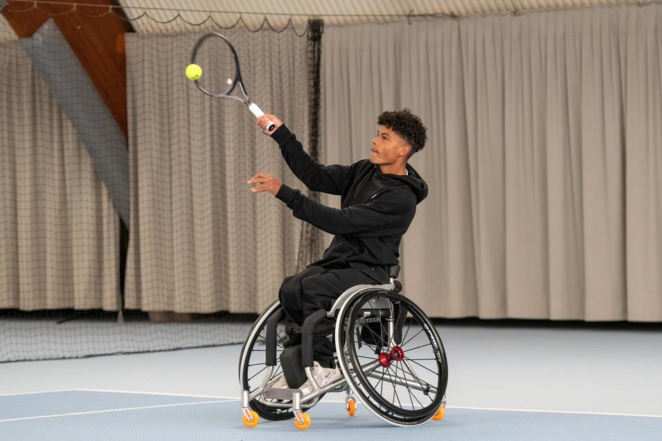 Junger Mann im Rollstuhl schlägt mit einem Tennisschläger einen Ball auf einem Indoor-Tennisplatz.