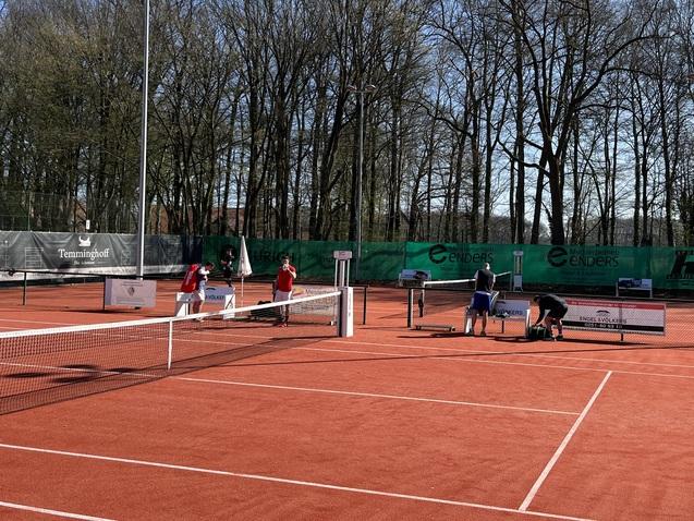 Tennisplatz mit mehreren Spielern in Aktion und Bäume im Hintergrund, bei sonnigem Wetter auf rotem Sand.
