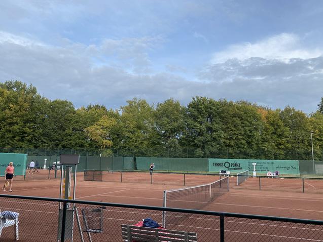 Tennisplatz mit zwei Spielern beim Spiel, umgeben von grünen Bäumen und bewölktem Himmel. Sitzbank im Vordergrund.