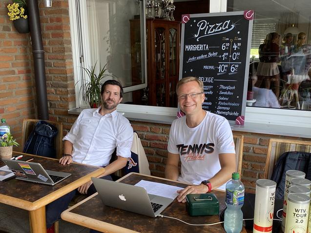 Zwei Männer sitzen an Tischen im Freien, arbeiten an Laptops und haben Getränke vor sich. Im Hintergrund steht eine Speisekarte.