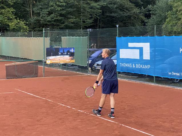 Tennis-Spieler auf einem roten Platz, bereit zum Aufschlag, umgeben von grünen Bäumen und einem Werbebanner.