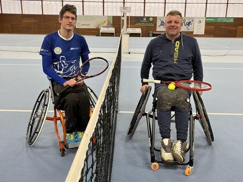 Zwei Rollstuhltennisspieler mit Schlägern und einem Ball sitzen auf gegenüberliegenden Seiten des Netzes in einem Tennisplatz.