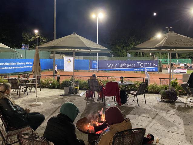 Tennisplatz bei Nacht mit Spielern, überdachten Sitzbereichen und einer Feuerschale im Vordergrund. Beleuchtung und Banner sichtbar.